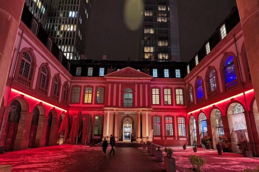 Ein historisches Gebäude mit Rundbogenfenstern und einem zentralen Eingang, beleuchtet durch rote Beleuchtung. 3
