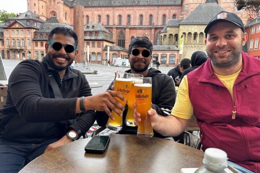 Three people toasting with beer glasses at a table in a public square with historic buildings in the background. 3