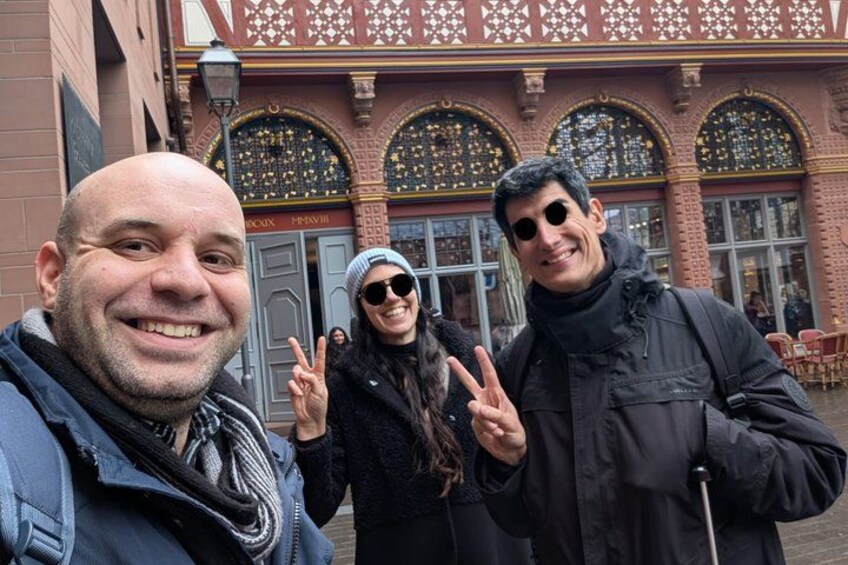 Tres personas hacen el signo de la paz frente a un edificio con ventanas ornamentadas y un letrero que dice 'BLOCH'. 3