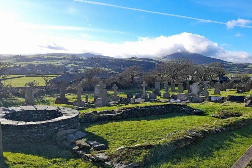 A cemetery with stone crosses and a circular stone structure, surrounded by green fields and a mountain in the background. 5
