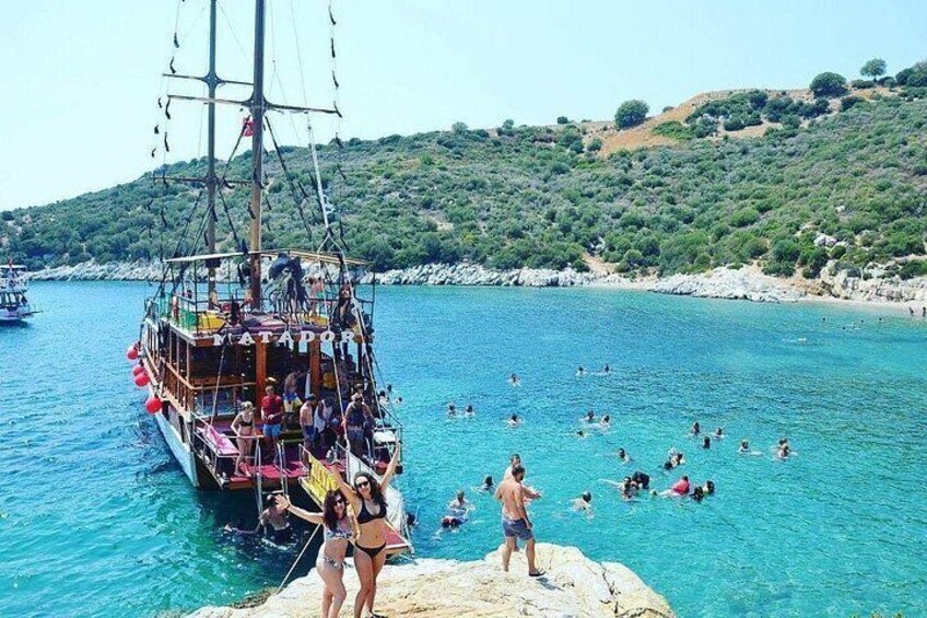 Un bote con gente, una orilla rocosa con nadadores y un paisaje montañoso al fondo. 1