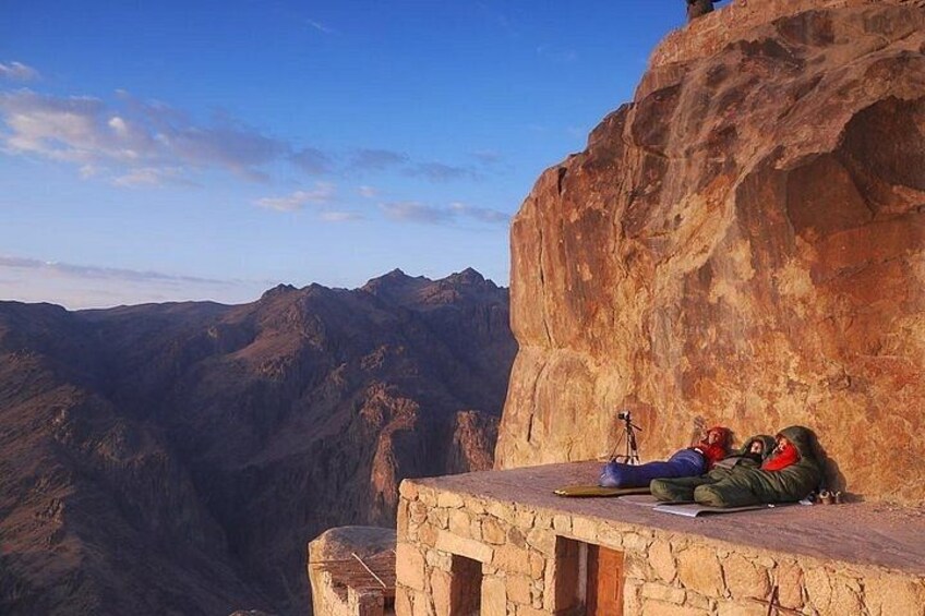A rocky cliff with a person lying on the edge, a backpack, and a camera on a tripod. 2