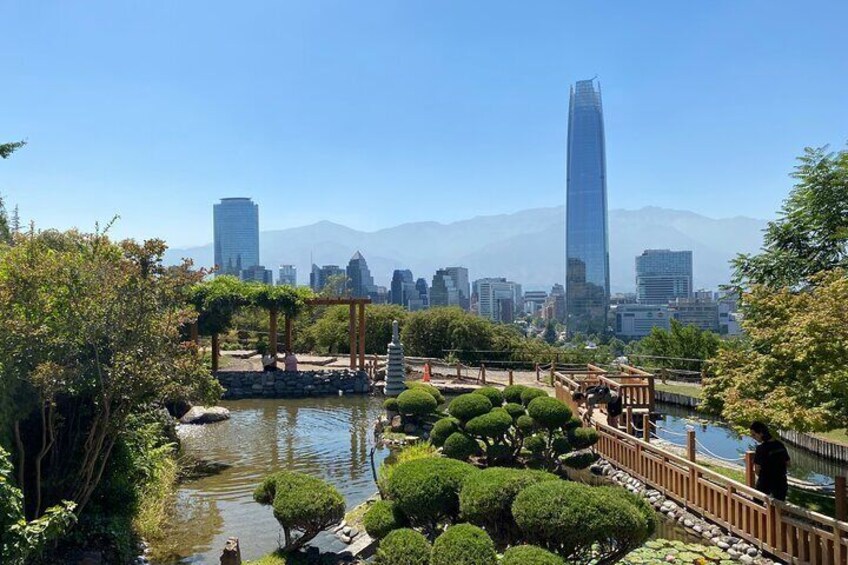 A park with a pond, neatly trimmed bushes, a wooden bridge, and a city skyline in the background. 1