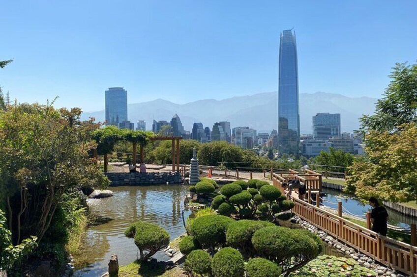 A serene park with a pond, neatly trimmed bushes, a wooden bridge, and a city skyline in the background. 2