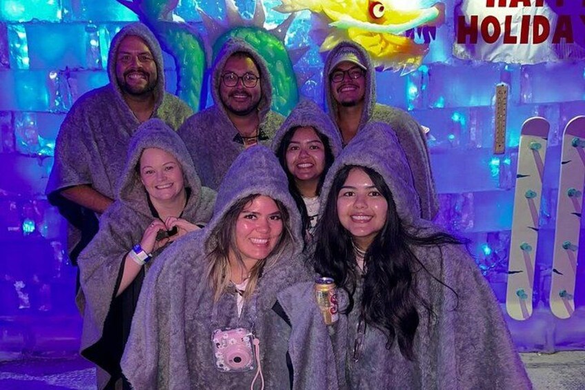 A group of people in winter clothing posing in front of an ice sculpture with a 'Merry Christmas' sign. 4