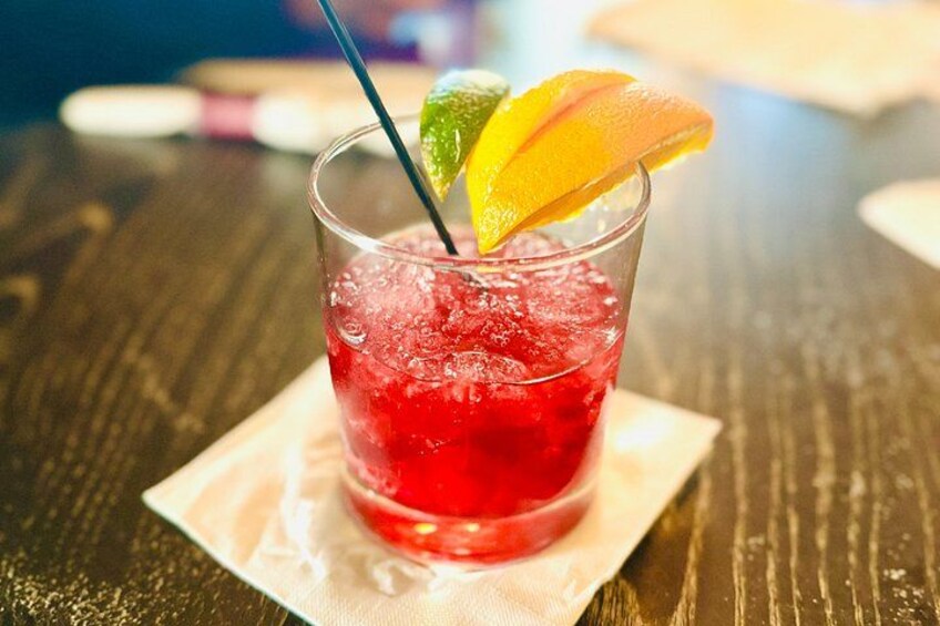 A glass of red beverage with ice cubes, garnished with a lime wedge and an orange slice, on a wooden table. 2