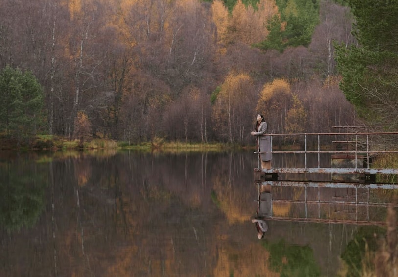 A person standing on a dock by a calm lake with autumn-colored trees in the background. 3