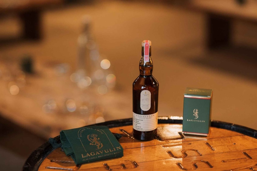 A bottle of Lagavulin whisky, a box of Lagavulin whisky, and a green booklet on a wooden barrel. 2