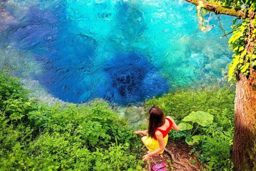 A person in a red top and yellow skirt standing near a vibrant blue water body surrounded by lush greenery. 5