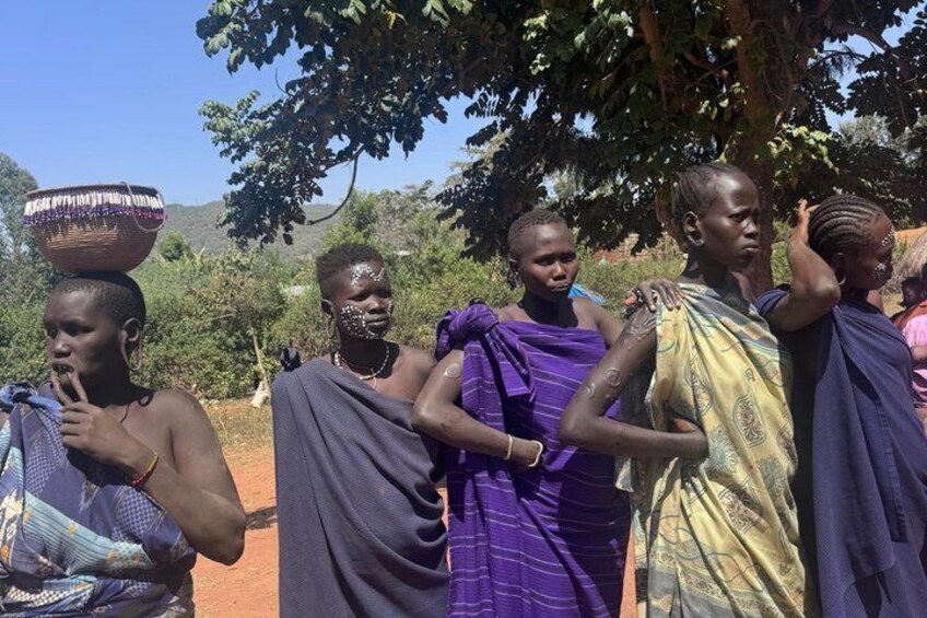 A group of people in traditional attire, some carrying baskets, standing outdoors with trees and hills in the background. 4