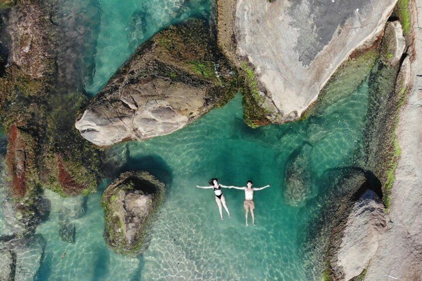 Two people swimming in clear turquoise water surrounded by large rocks and boulders. 2