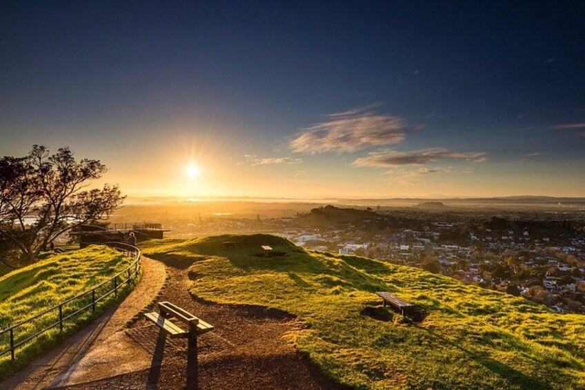 A sunset over a grassy hill with a path leading to a bench, overlooking a cityscape. 2