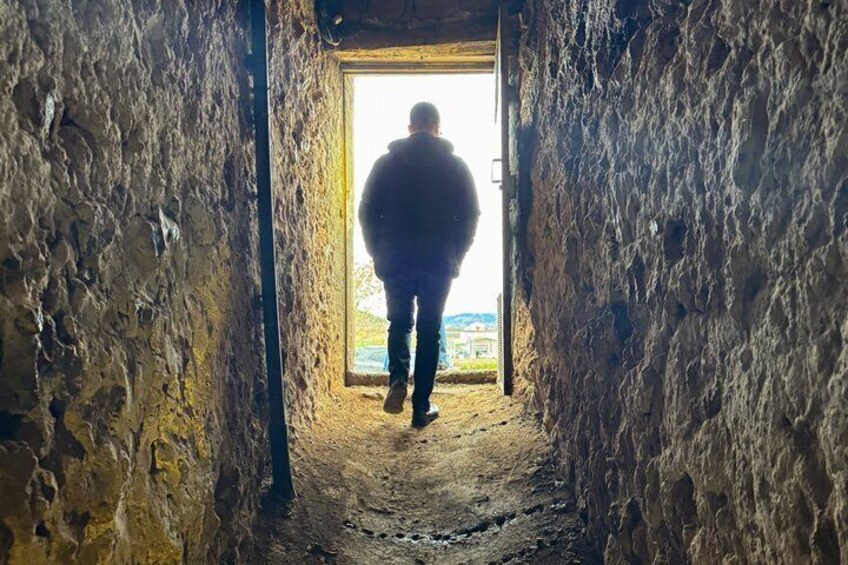 Eine Person geht durch einen schmalen, steinernen Gang mit rauen Wänden und einem Durchgang zu einem offenen Bereich. 5