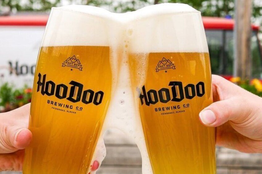 Two glass mugs with 'Hoodoo Brewing Co' branding, held by a person. 1