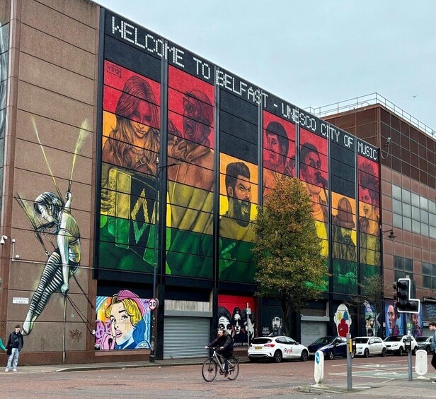 A building with a large mural welcoming visitors to Belfast, featuring various portraits and a street artist's sculpture. 2