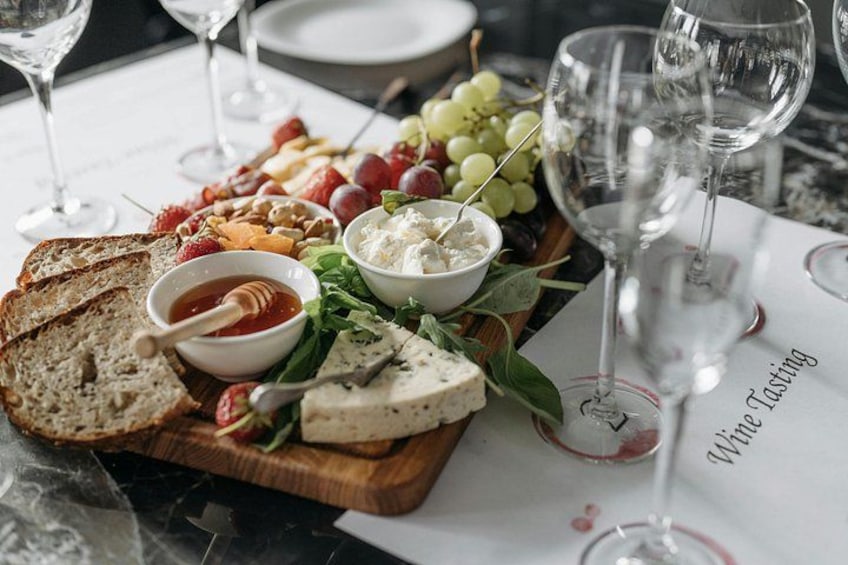 A charcuterie board with cheese, fruits, and bread on a wooden board, surrounded by wine glasses on a table. 4