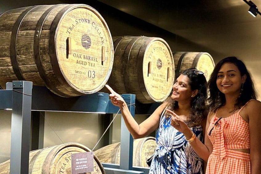 Zwei Personen zeigen auf Holzbottiche mit dem Aufdruck 'OAK BARREL AGED TEA' in einem Lagerraum. 4