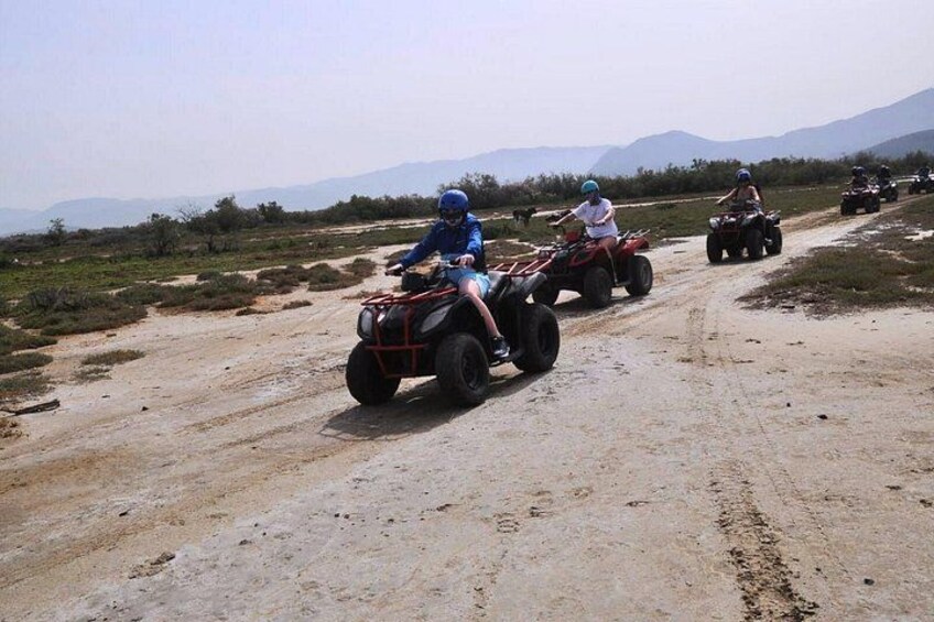 A group of people riding ATVs on a dirt road in a rural area with sparse vegetation and mountains in the background. 3