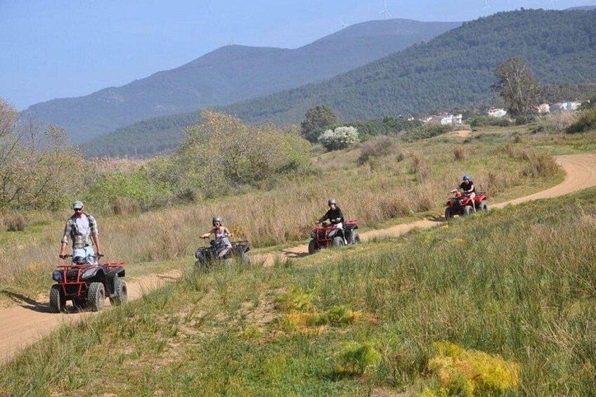 Four people on quad bikes on a dirt path with grass and trees, surrounded by hills and houses in the background. 5