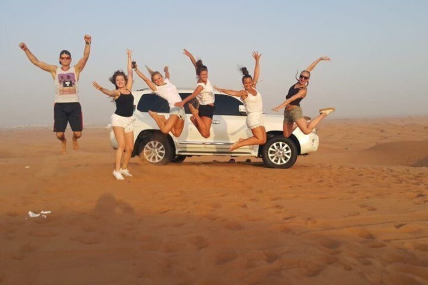 A group of people jumping in the desert near a white vehicle. 2
