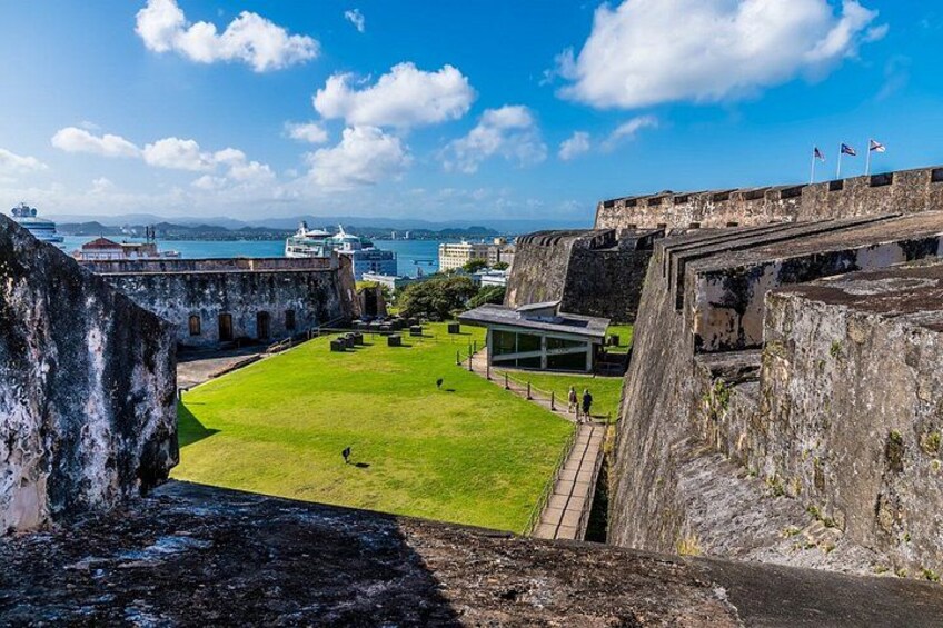 A historic fort with a grassy courtyard, a modern building, and a view of the sea with cruise ships. 5