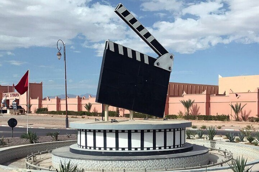 Une claquette de cinéma posée sur une plateforme circulaire, avec un paysage désertique et des bâtiments en arrière-plan. 3