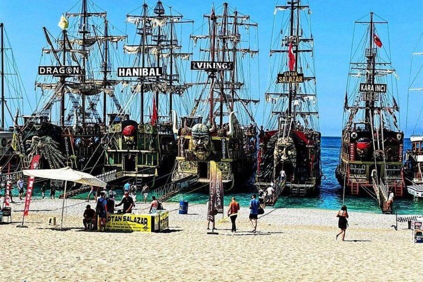A beach with several large, ornate sailing ships docked. 5