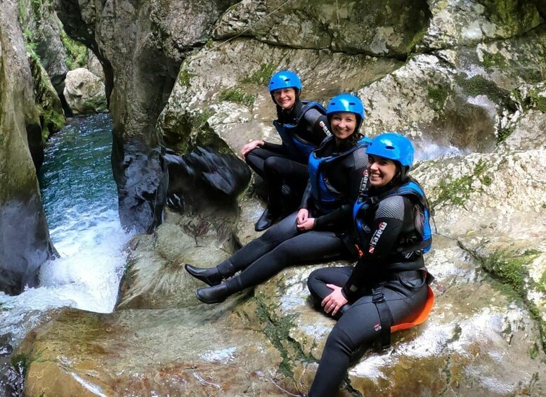 Two people in black wetsuits and blue helmets sitting on a rock by a waterfall. 4