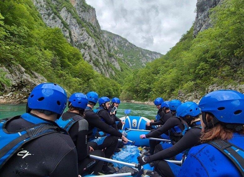 A group of people in blue helmets and wetsuits on a white raft, paddling through a river surrounded by green cliffs. 1