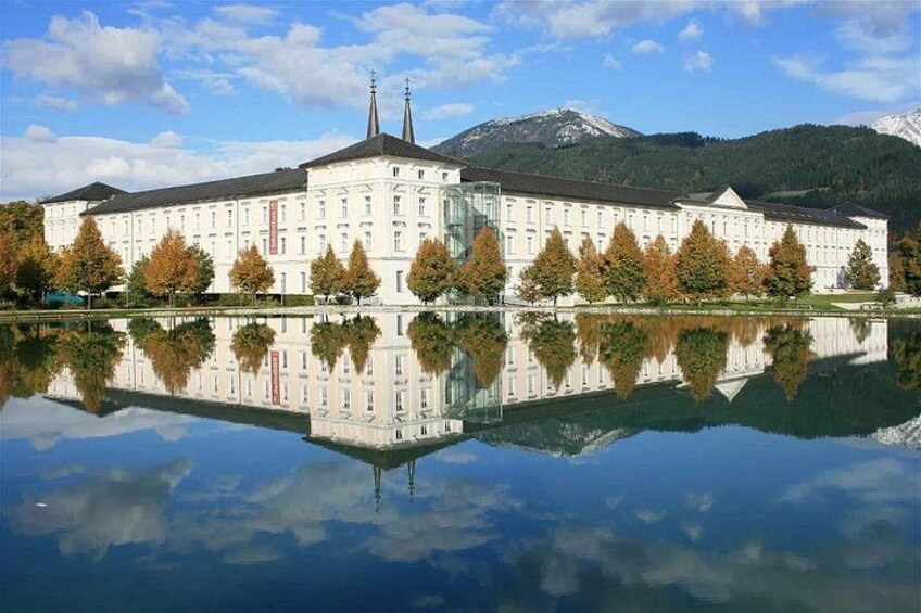 A large white building with a reflection in a calm body of water, surrounded by autumn trees and a mountain in the background. 2