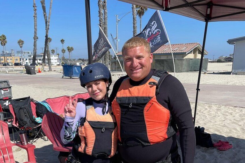 Two people wearing life jackets and helmets, a beach, palm trees, and a flag. 1