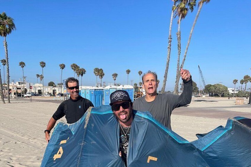 Tre personer bærer et stort, blåt telt på en strand med palmer og mobile toiletter i baggrunden. 1