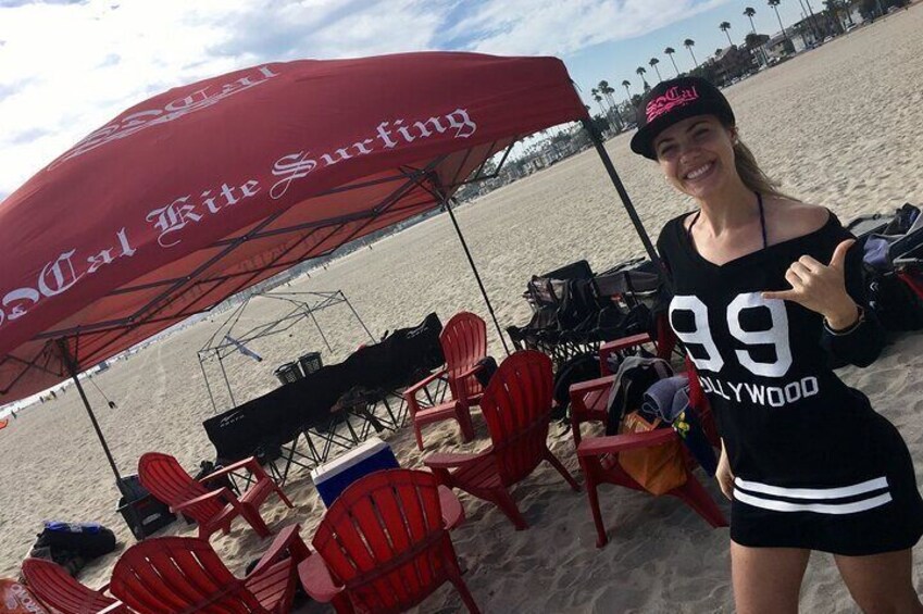 En person står under en rød parasol med teksten 'Cal Kite Surfing', på en sandstrand med røde stole og en sort køleboks. 2