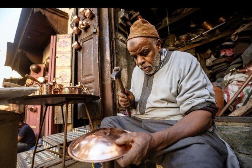 A man sitting at a table with brass items, possibly a workshop or market. 4