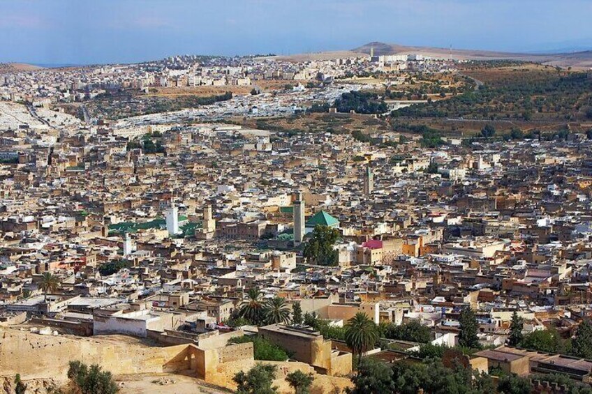 A densely populated cityscape with numerous buildings, a prominent mosque with a green dome, and a hill in the background. 1