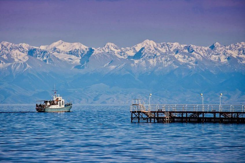 A boat on calm water, a pier extending into the water, and snow-capped mountains in the background. 1