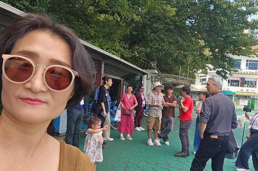 A group of people gathered outdoors, some standing and conversing, with trees and a building in the background. 3