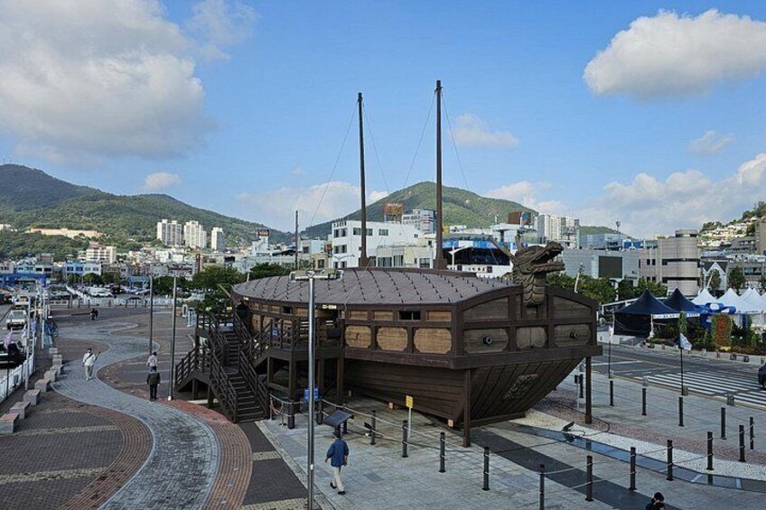 A wooden ship-shaped building with stairs, a pedestrian walkway, and a mountainous backdrop. 1