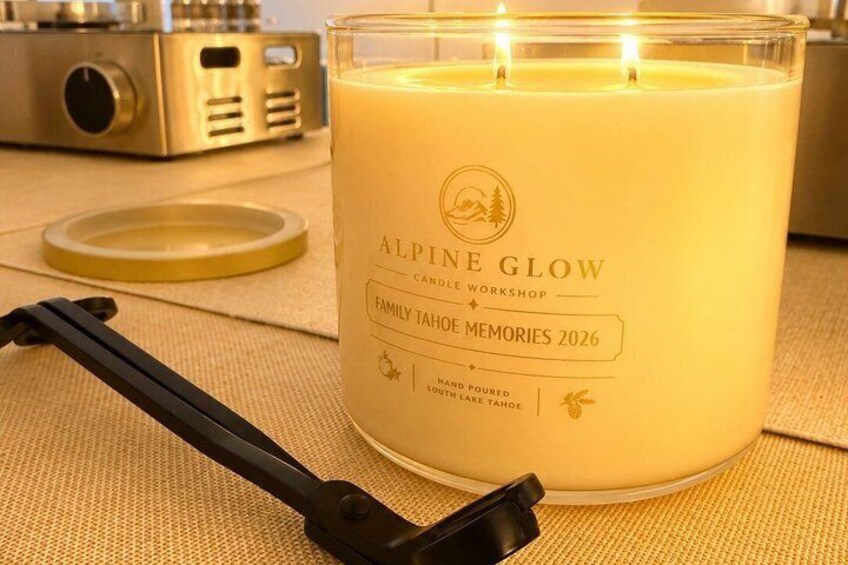 A lit candle labeled 'Alpine Glow Candle Workshop' with a 'Family Tahoe Memories 2026' design, placed on a textured surface with a black candle snuffer nearby. 1