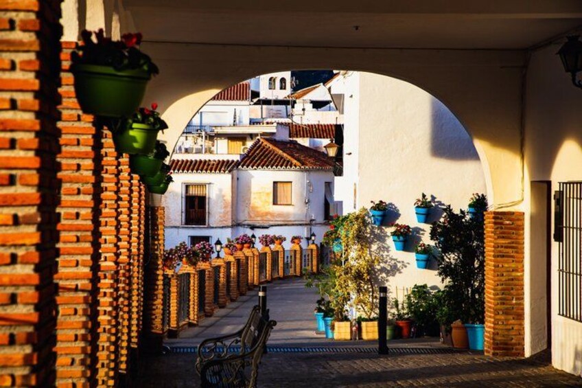 A narrow alleyway with brick walls, hanging flower pots, and a bench. 1