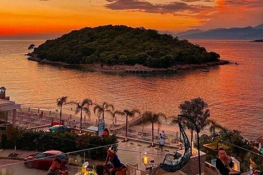 A sunset over a body of water with a small island, a beach with umbrellas, and a dining area with people. 3
