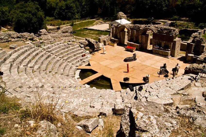 An ancient amphitheater with stone seating and a central stage, surrounded by ruins and greenery. 1