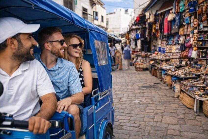 A blue tuk-tuk with passengers, a cobblestone street, and a market with various goods. 5