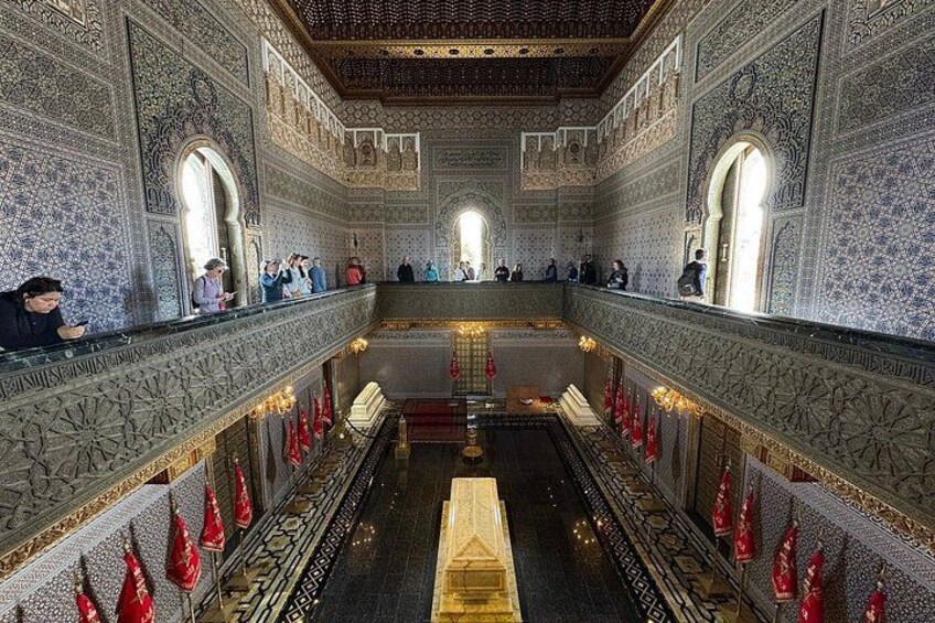 An ornate interior with intricate tile work, a central golden structure, and red flags lining the walls. 5