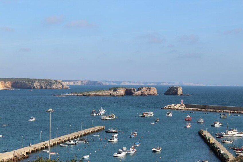 A coastal marina with boats docked at piers, rocky cliffs in the background, and a lighthouse on a small island. 2