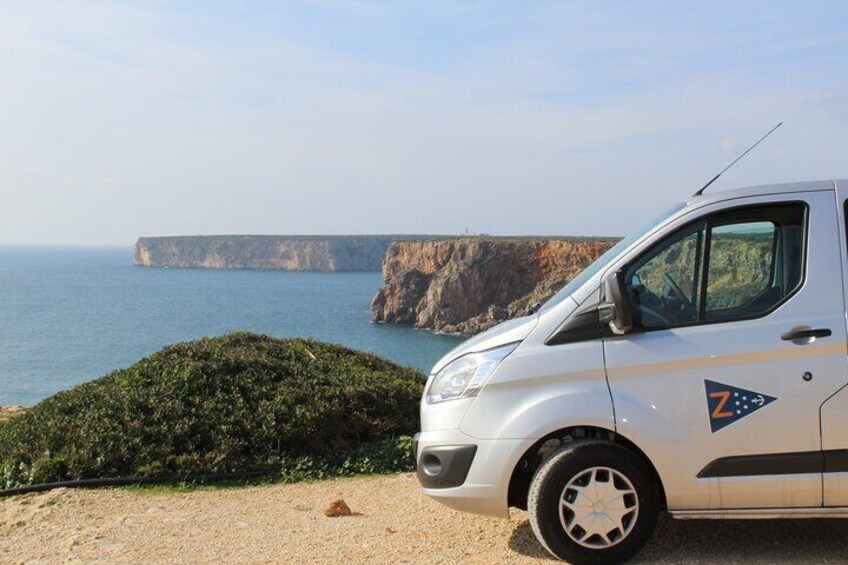 A white van parked on a dirt path with a cliff and ocean in the background. 5