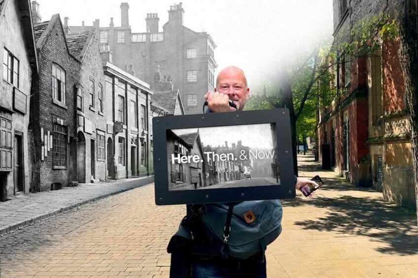 Eine Person hält ein gerahmtes Bild mit dem Text 'Here. Then. & Now.' in einer Straße mit alten Gebäuden. 1