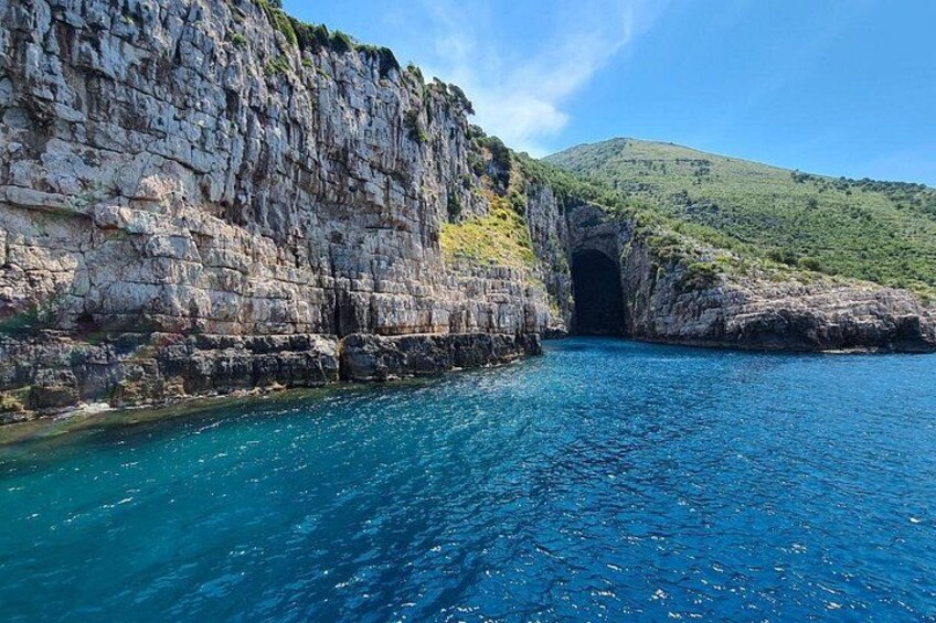 A coastal landscape with a rocky cliff, a clear blue sea, and a natural cave entrance. 2