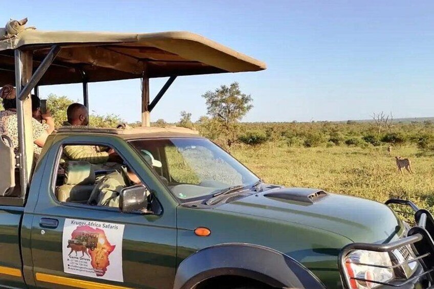 A safari vehicle with a sign for Kruger Africa Safaris, a person sitting inside, and a landscape with sparse vegetation and a distant animal. 3