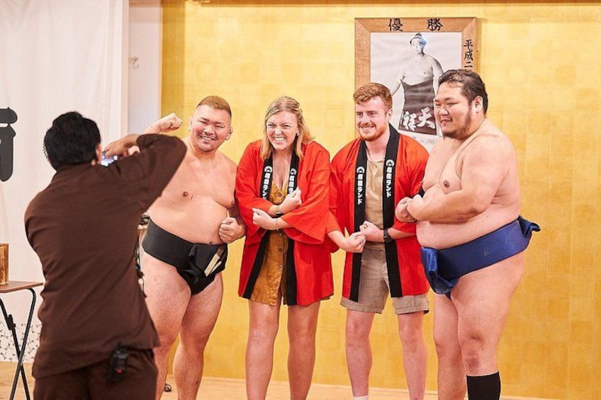 En sumobrottningsmatch med deltagare i traditionella dräkter, en fotograf som dokumenterar evenemanget och en affisch i bakgrunden. 2
